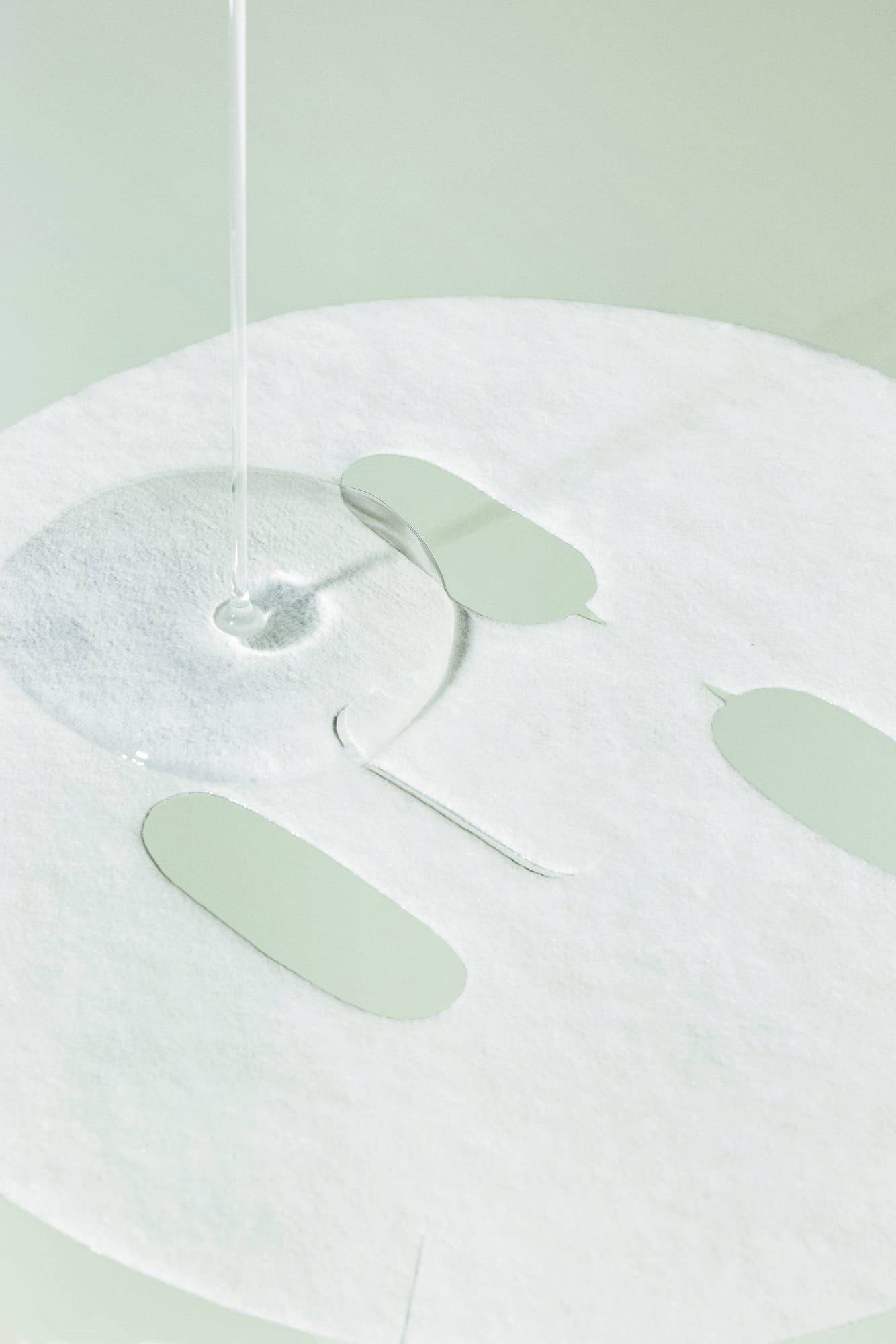 A close-up of a white sheet mask with cutout eye, nose, and mouth shapes, with a clear liquid being poured over the surface against a pale mint green background.