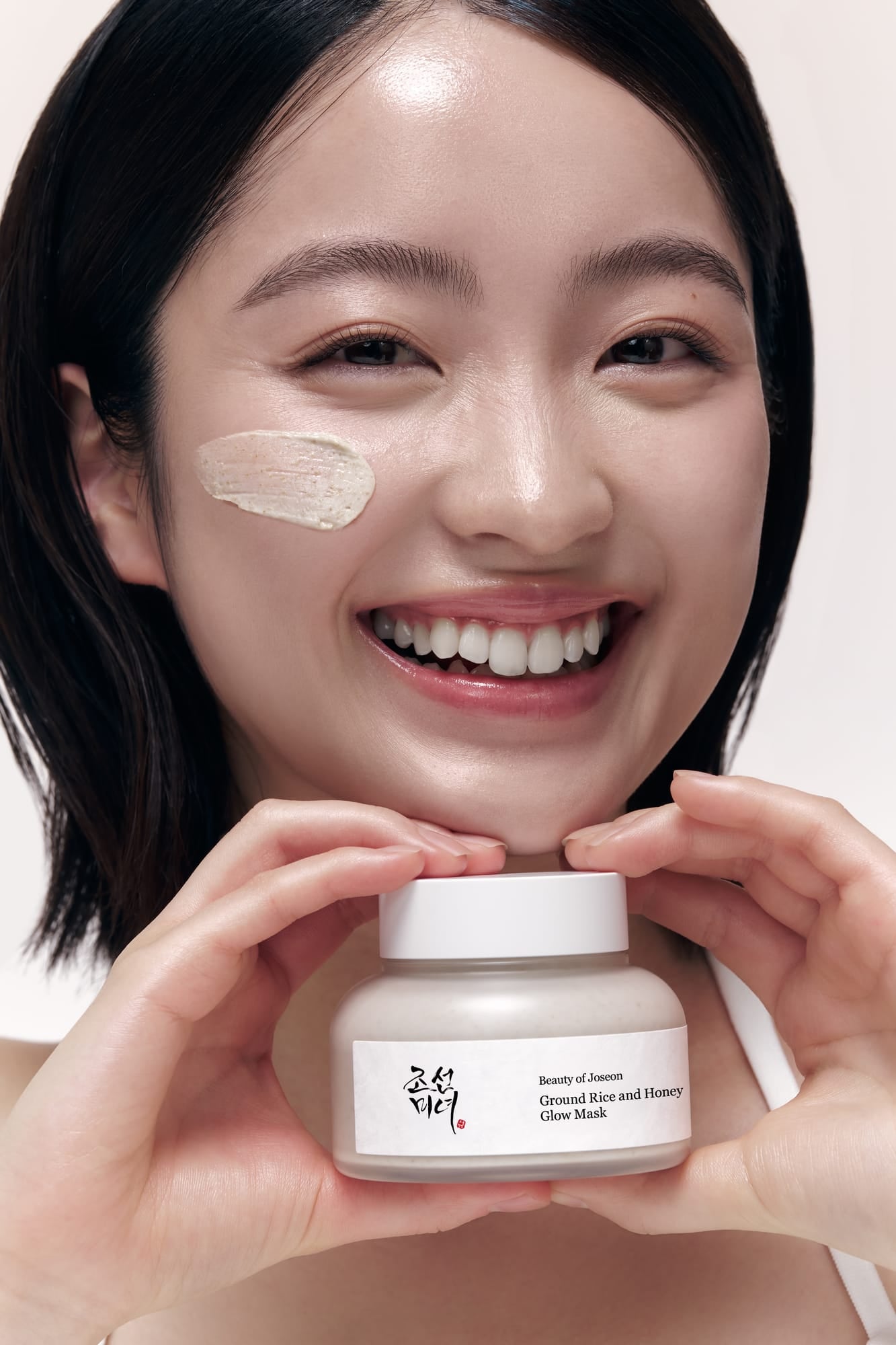 A smiling woman with dark hair holds a jar of glow mask in front of her face, with a dollop of the cream-colored mask on her cheek. The product label reads Beauty of Joseon Glow of Beauty Covered Skin and History Glow Mask.